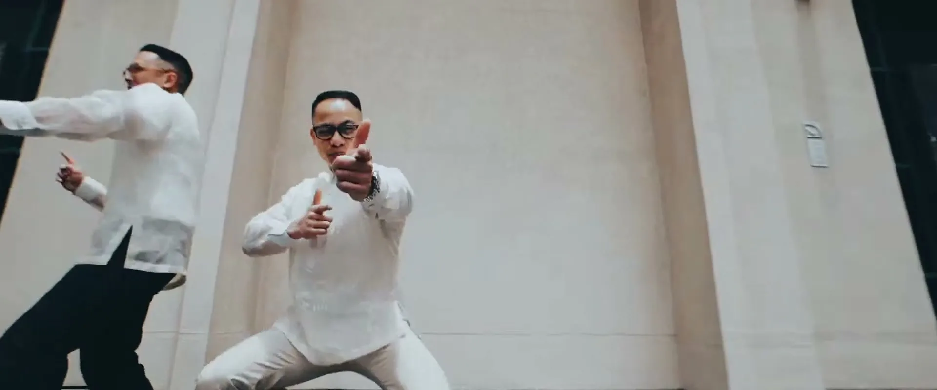 Two men in white shirts and dark pants perform a synchronized dance routine against a plain wall, with one man pointing at the camera.