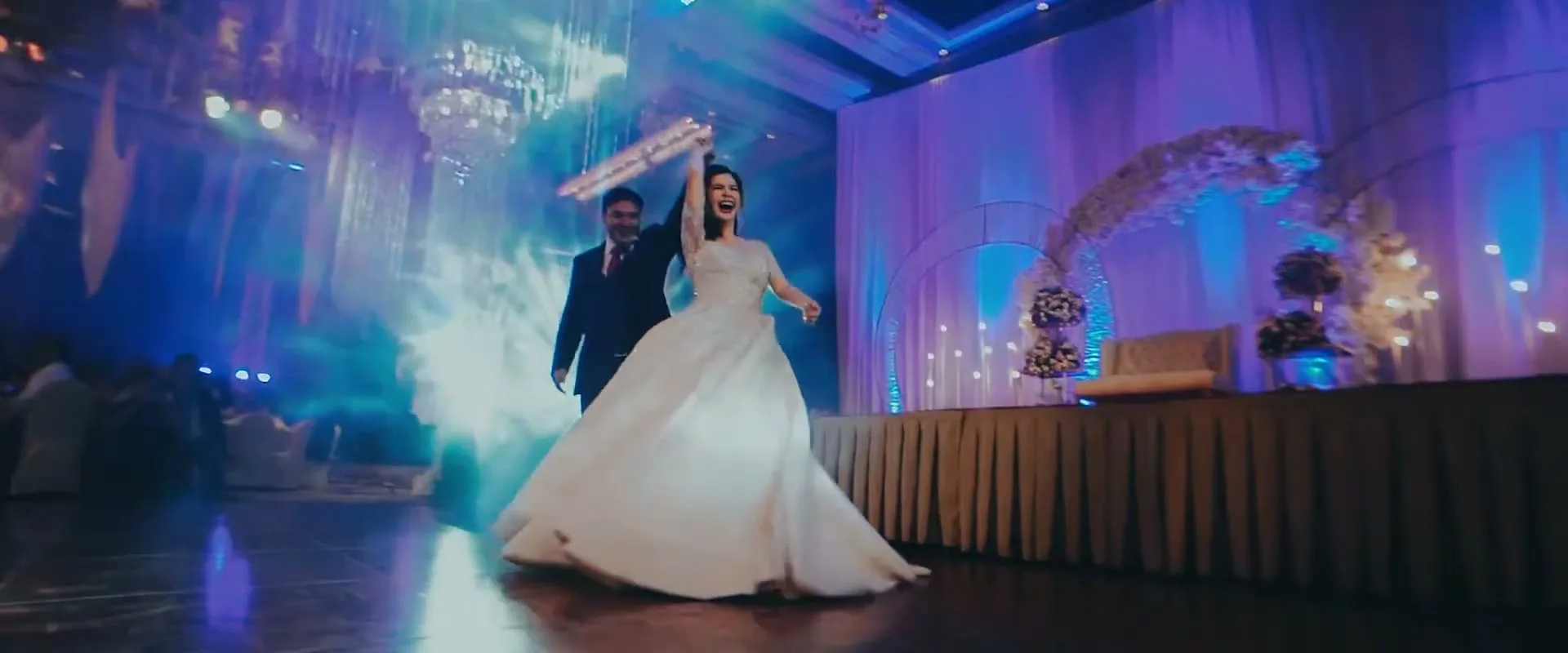 A joyful bride in a white gown dances with her groom in a dark suit at their wedding reception, surrounded by purple and blue lights.