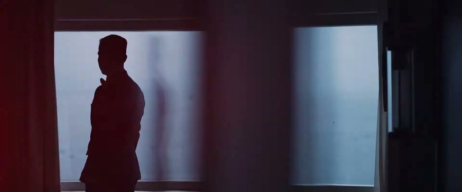 A silhouetted man in a suit stands by a window, looking out, in a dimly lit room.