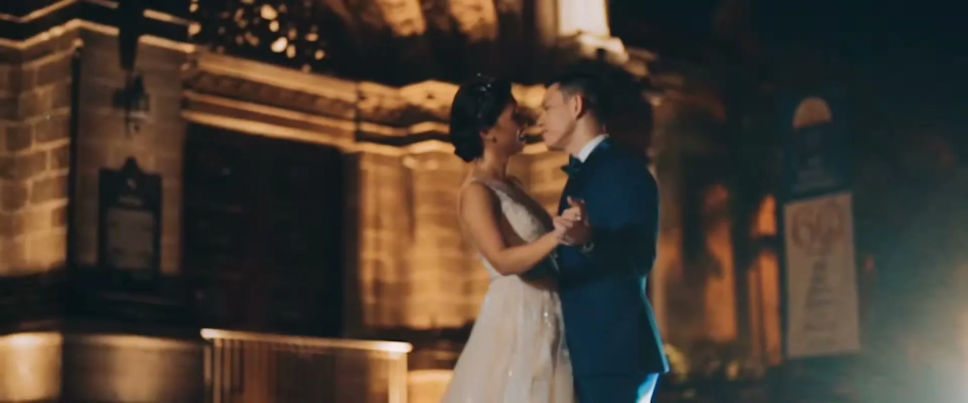 A newlywed couple, a woman in a white wedding dress and a man in a blue suit, dance together at night in front of an old stone building.