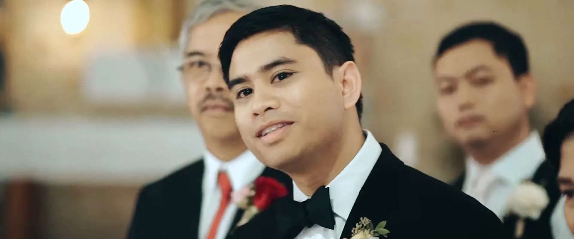 A groom in a black tuxedo with a bow tie and boutonniere looks up and smiles during a wedding ceremony inside a church.