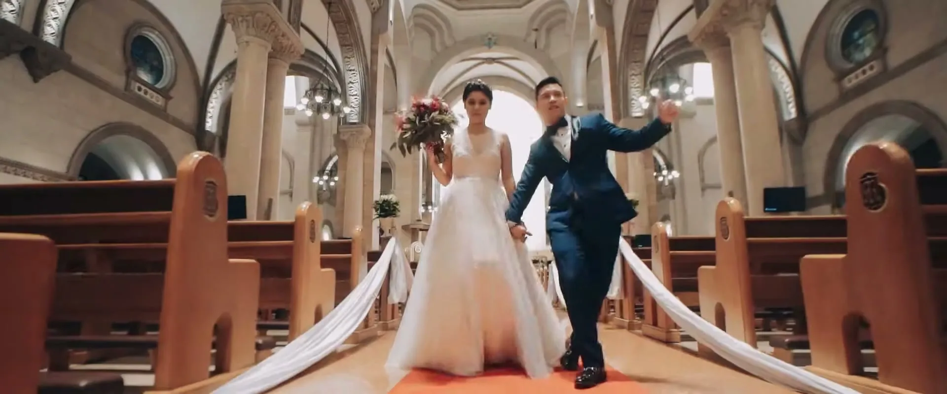 A newlywed couple, a woman in a white wedding dress and a man in a blue suit, walk hand-in-hand down the aisle of a church, smiling and gesturing.