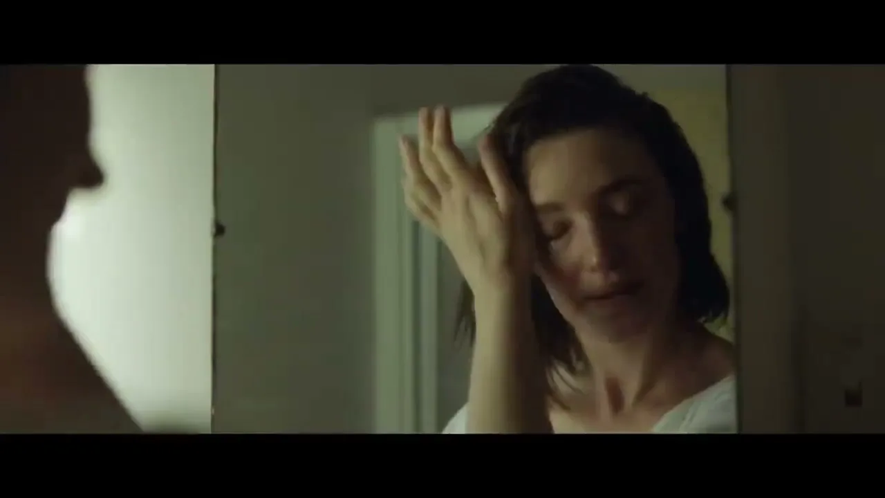 A woman with short dark hair, wearing a white top, looks at her reflection in a mirror, touching her face with a sad expression in a dimly lit room.
