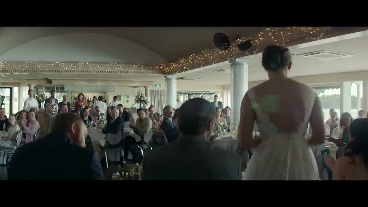 A wide shot shows a bride in a white dress standing with her back to the camera, facing a large group of wedding guests seated at tables in a bright reception hall.