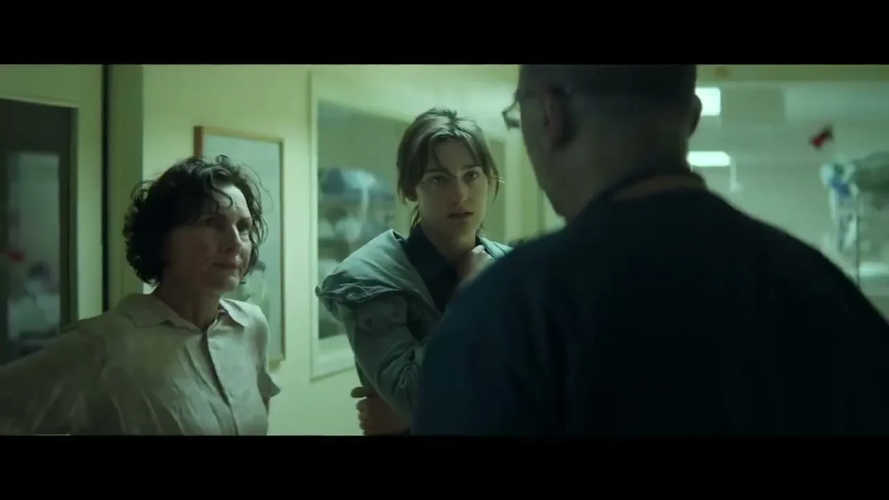 Two women, one with short curly hair and one with long brown hair, stand in a hospital hallway, looking concerned as they talk to a man whose back is to the camera.