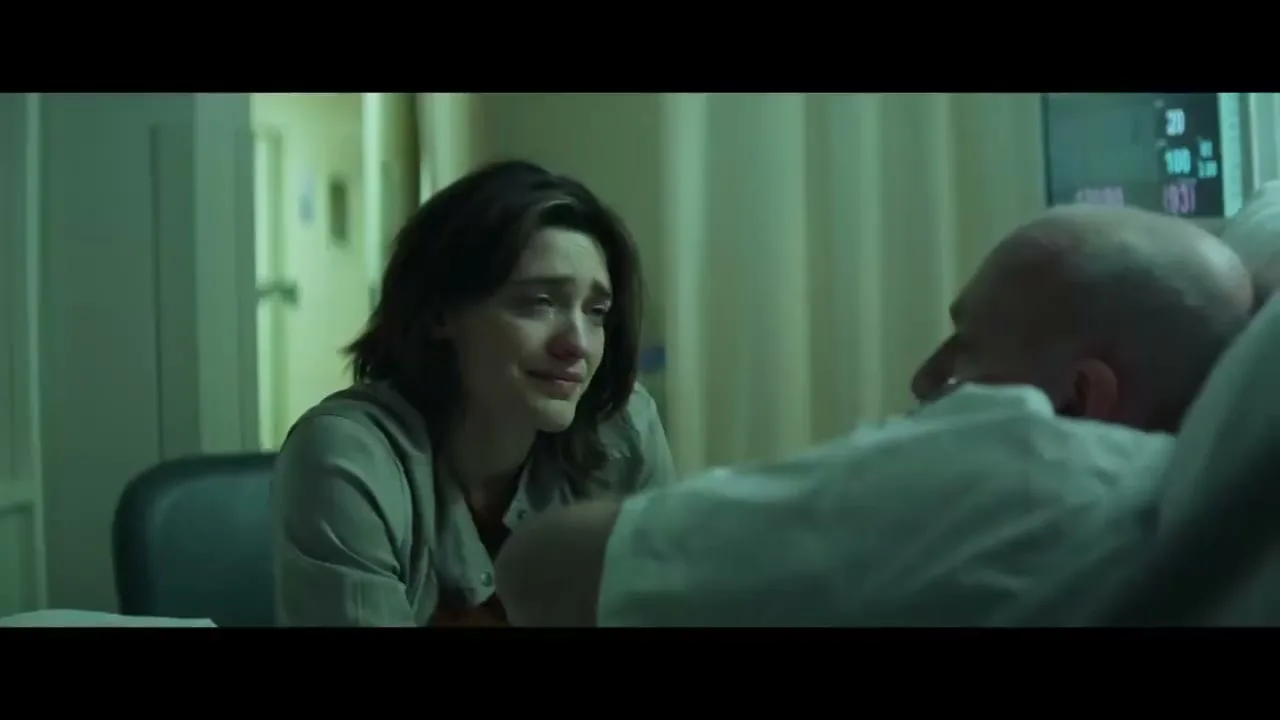 A young woman with dark hair, wearing a grey top, sits crying next to a bald man in a hospital bed, who is speaking to her.