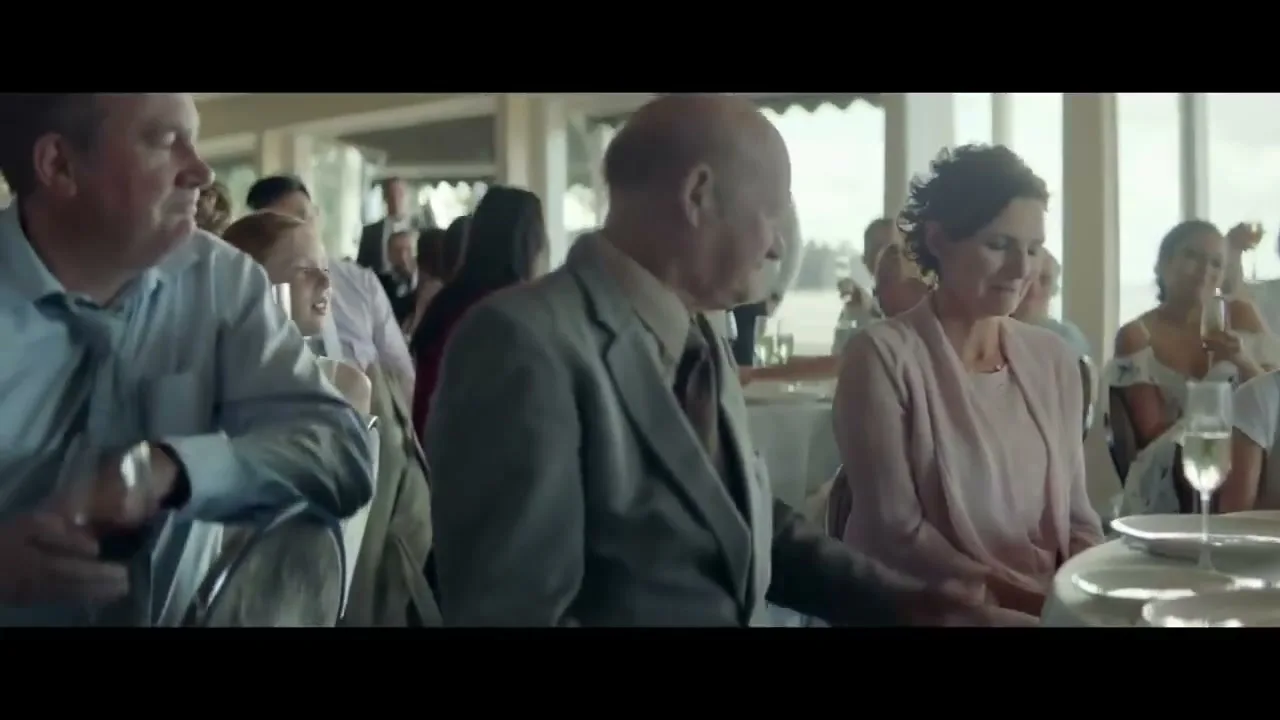 A diverse group of wedding guests, including a man in a blue shirt, an older man in a suit, and a woman in a pink dress, sit at tables in a bright, airy reception hall.