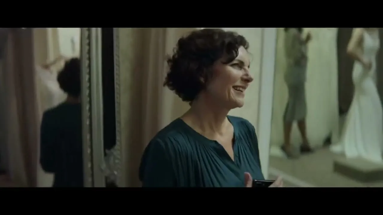 A woman with short, curly brown hair, wearing a dark teal top, smiles while holding a phone in a bridal shop.