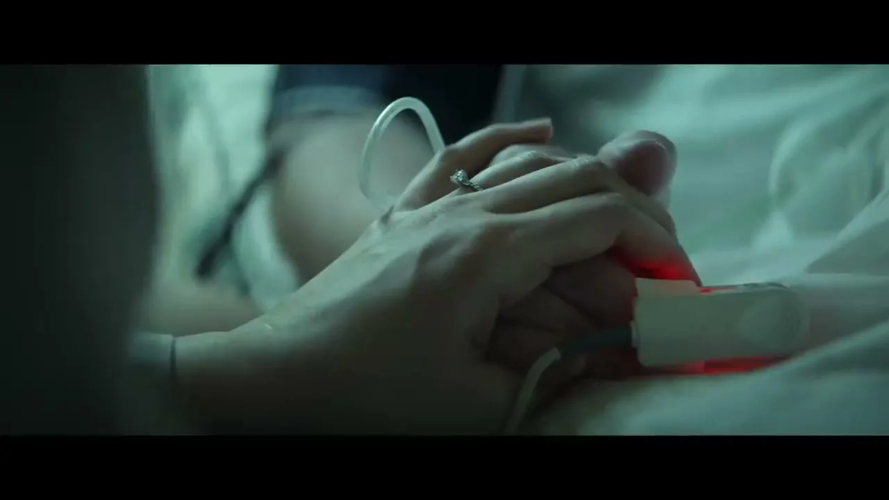 A woman's hand, wearing a diamond ring, holds a man's hand with a pulse oximeter on his finger, in a hospital bed.
