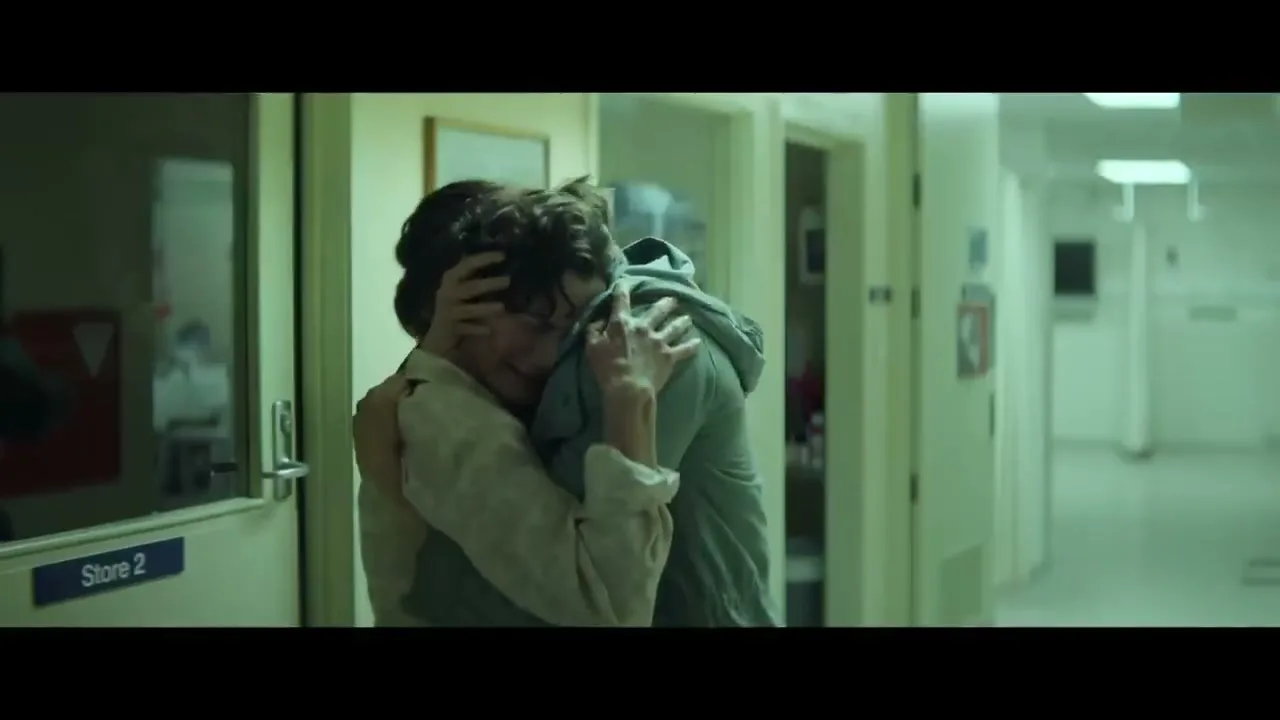 A young man with curly hair and a woman with long hair embrace tightly in a hospital hallway, both looking emotional.