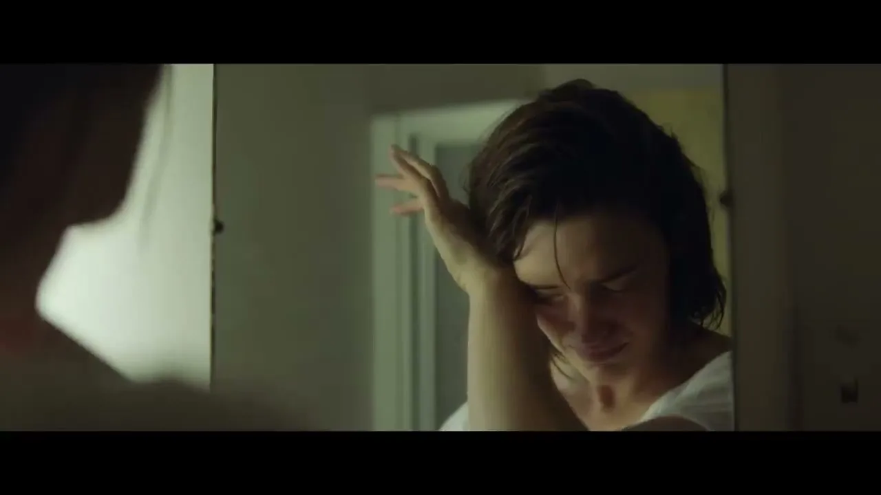 A woman with short brown hair, wearing a white t-shirt, looks at her reflection in a mirror, wiping her face and smiling in a dimly lit bathroom.