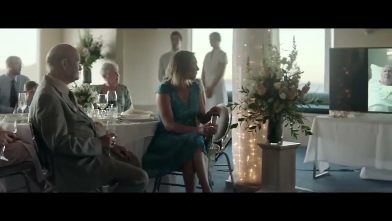 Wedding guests, including a man in a suit and a woman in a teal dress, sit at a table, watching a man on a TV screen at a wedding reception.