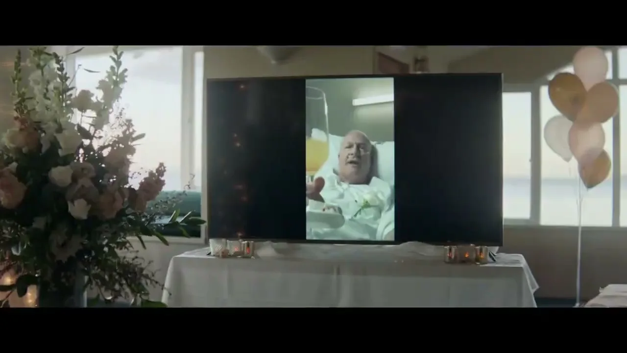 A wide shot shows a TV screen displaying a man in a hospital bed, raising a glass, flanked by flowers and balloons at a wedding reception.
