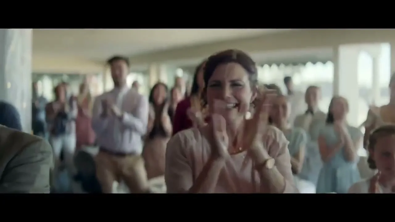 A woman in her 30s, wearing a light pink top, claps and smiles happily at a wedding reception, surrounded by other guests.