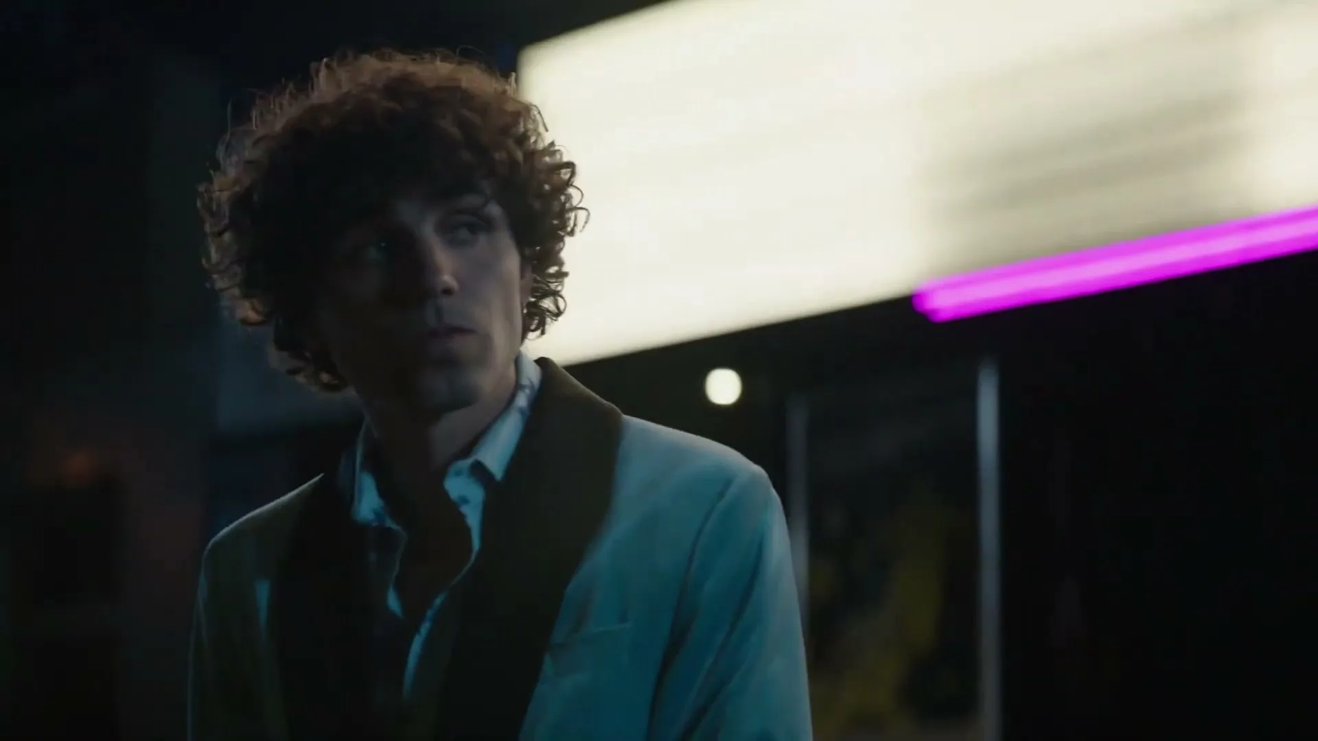 A young man with curly hair in a light suit looks around a dimly lit club or bar at night.