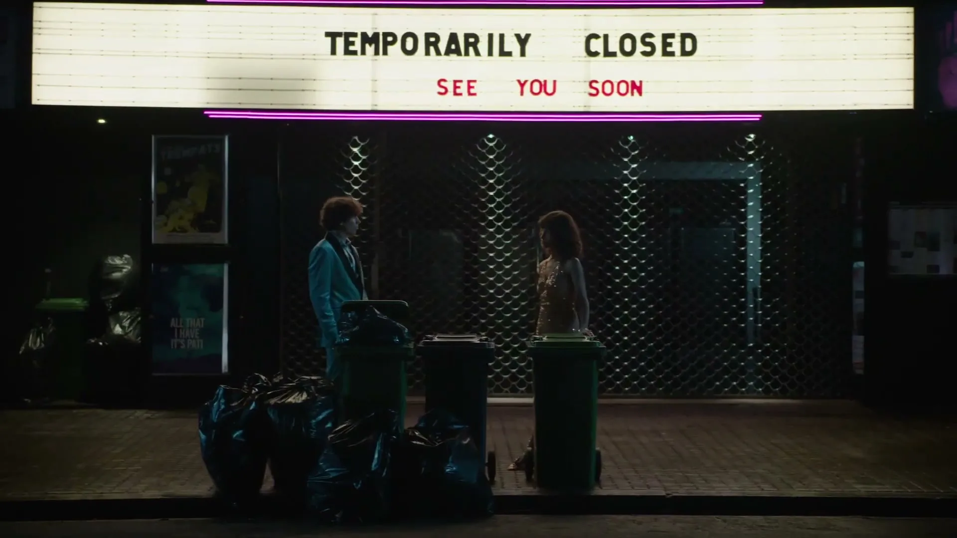 A young man and woman stand outside a closed theater at night, with trash bags and bins around them, under a sign that reads "TEMPORARILY CLOSED SEE YOU SOON."