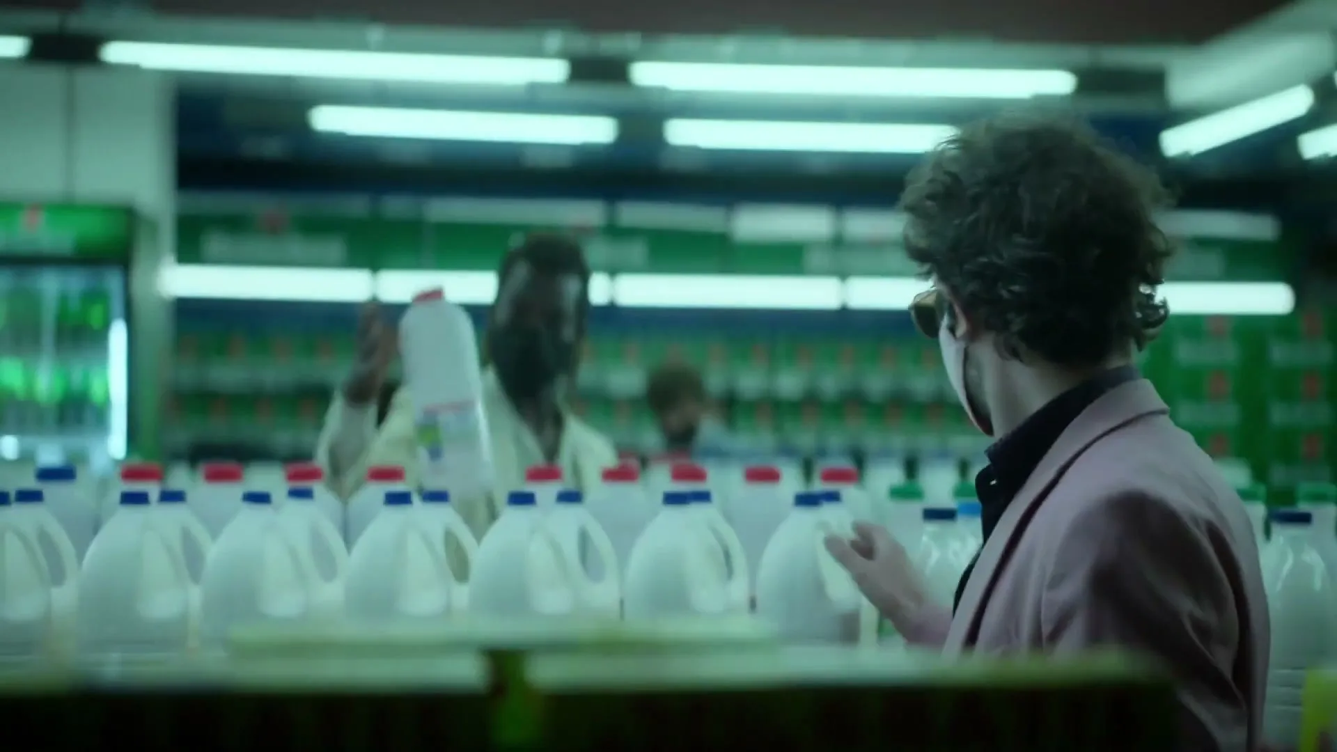 A man in a pink suit and face mask looks at milk bottles in a supermarket, then turns to see another man in a mask.
