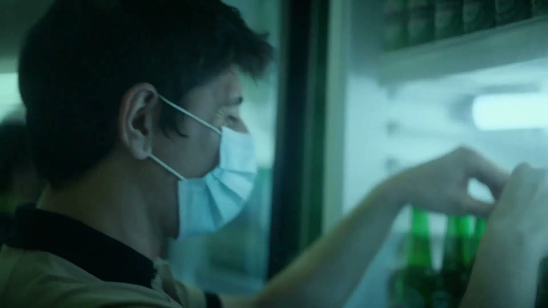 A man wearing a face mask looks over his shoulder while reaching into a refrigerator filled with Heineken bottles in a store.