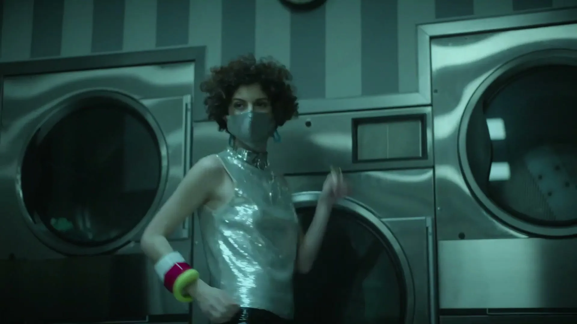 A woman with curly hair, wearing a sequined top and face mask, dances in a laundromat.