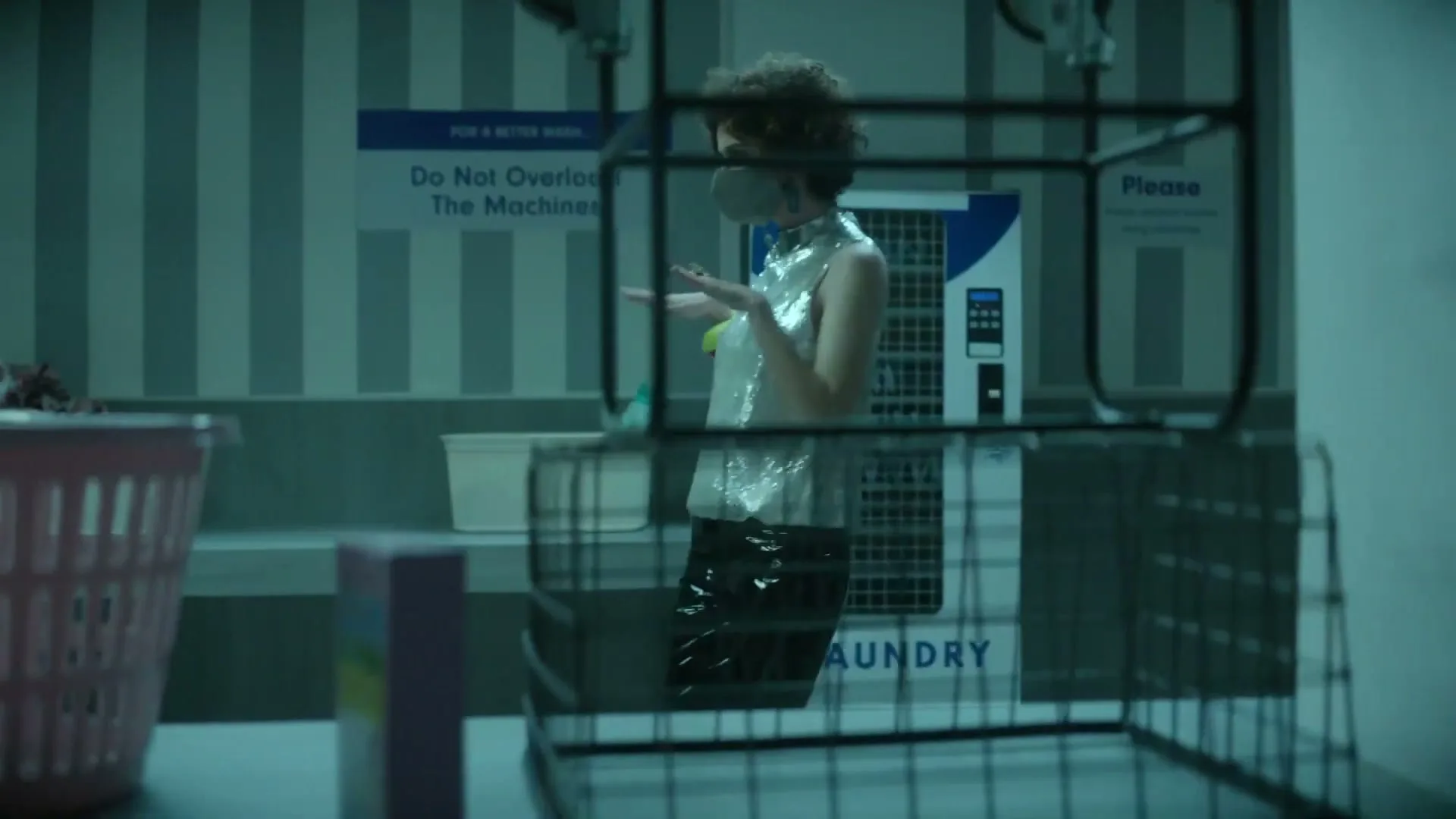 A young woman in a sparkly top and face mask stands in a laundromat, gesturing with her hands as if talking to someone off-screen.