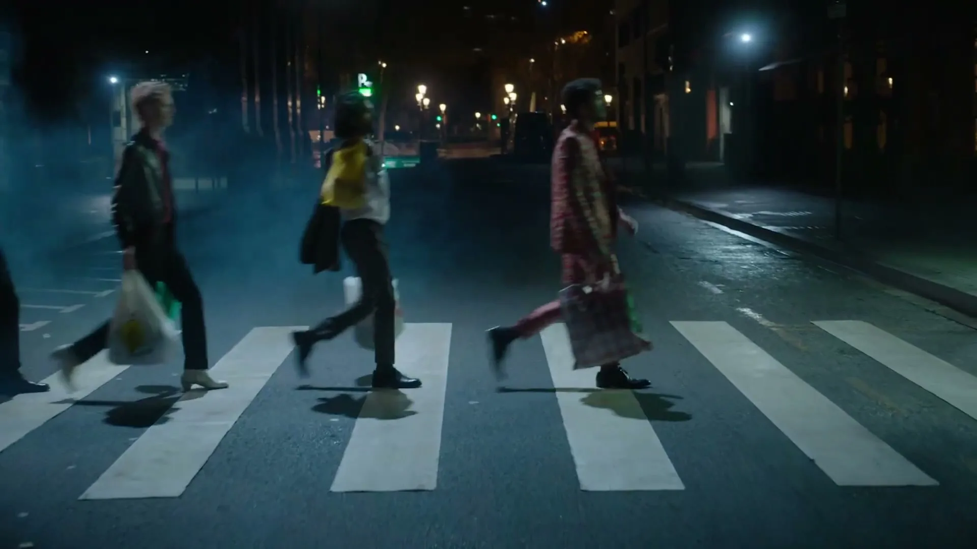 Four young adults, dressed in stylish, colorful outfits, walk across a zebra crossing on a city street at night, carrying shopping bags.