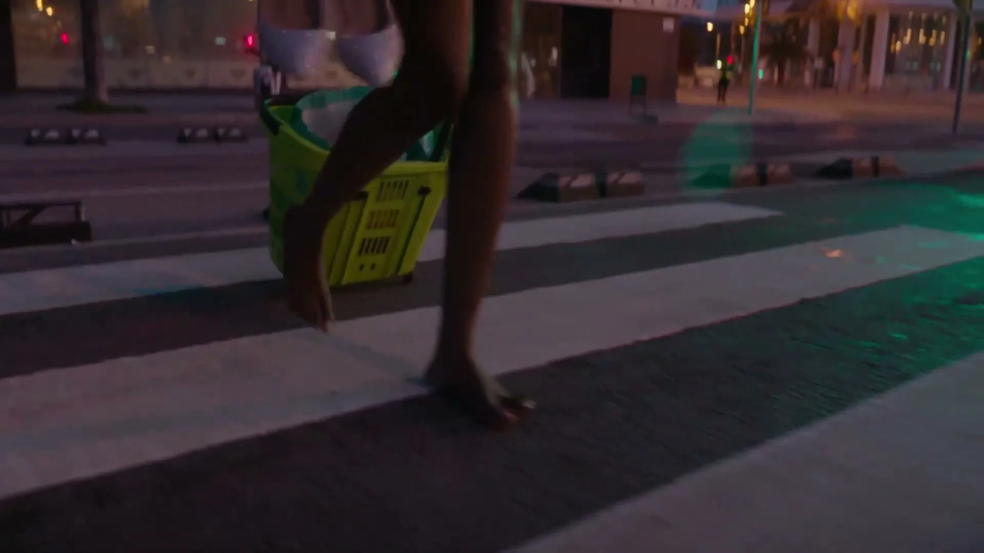 A person in a pink dress and carrying a green shopping basket walks barefoot across a crosswalk at dusk in a city.