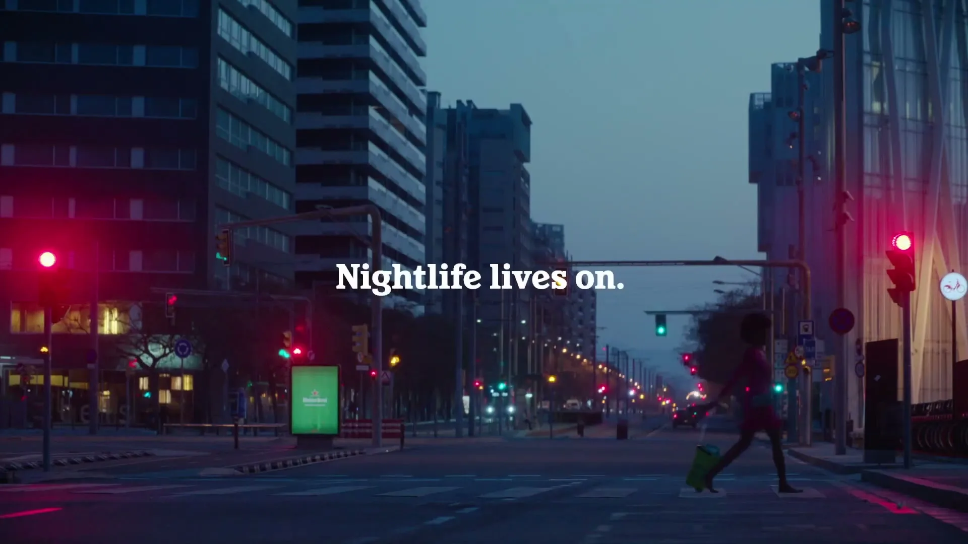 A woman in a pink dress and carrying a green suitcase crosses a wide, empty city street at dusk, with tall buildings and traffic lights in the background.