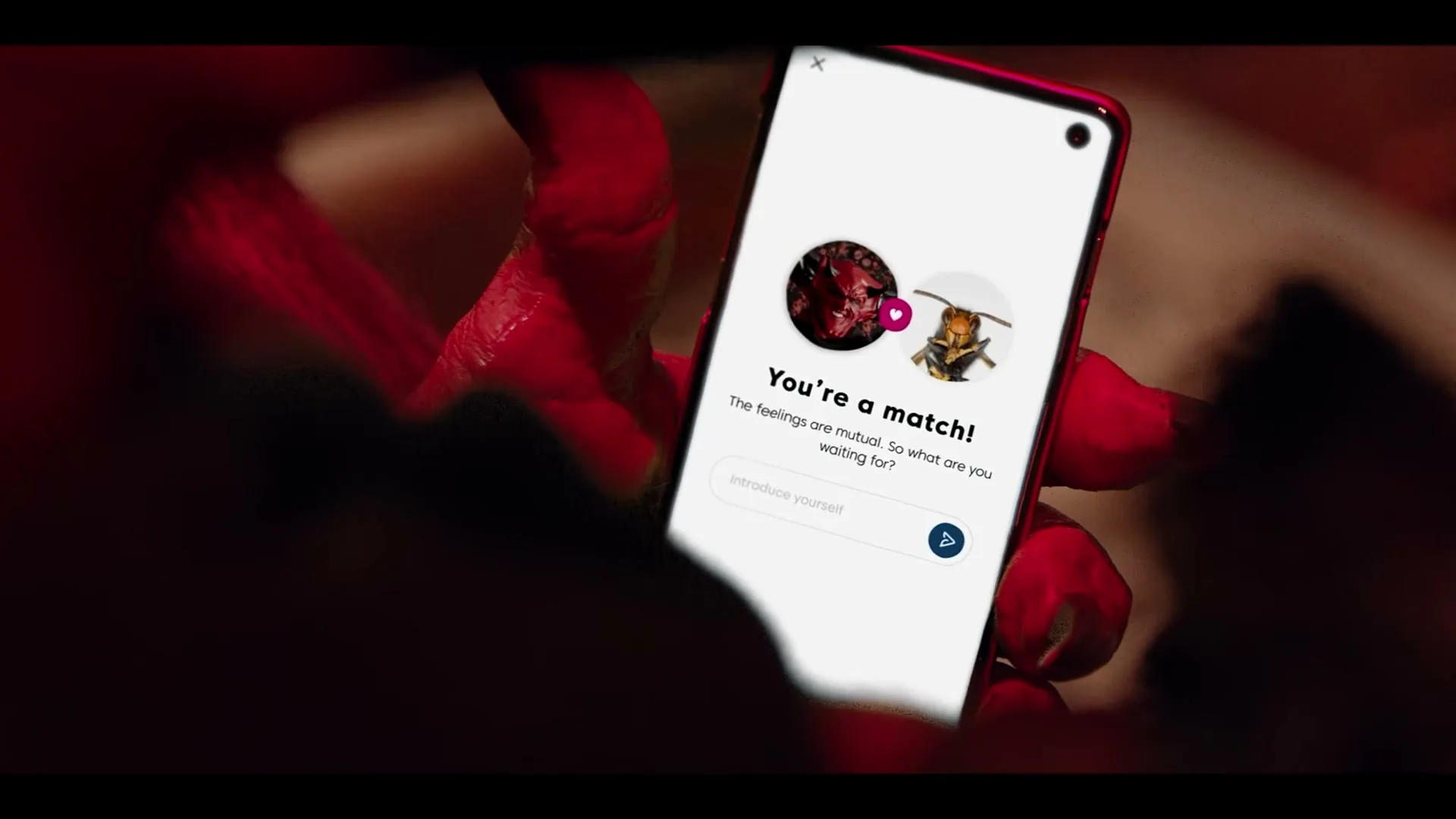 A close-up shot shows a red-skinned hand holding a smartphone displaying a dating app with a "You're a match!" notification.