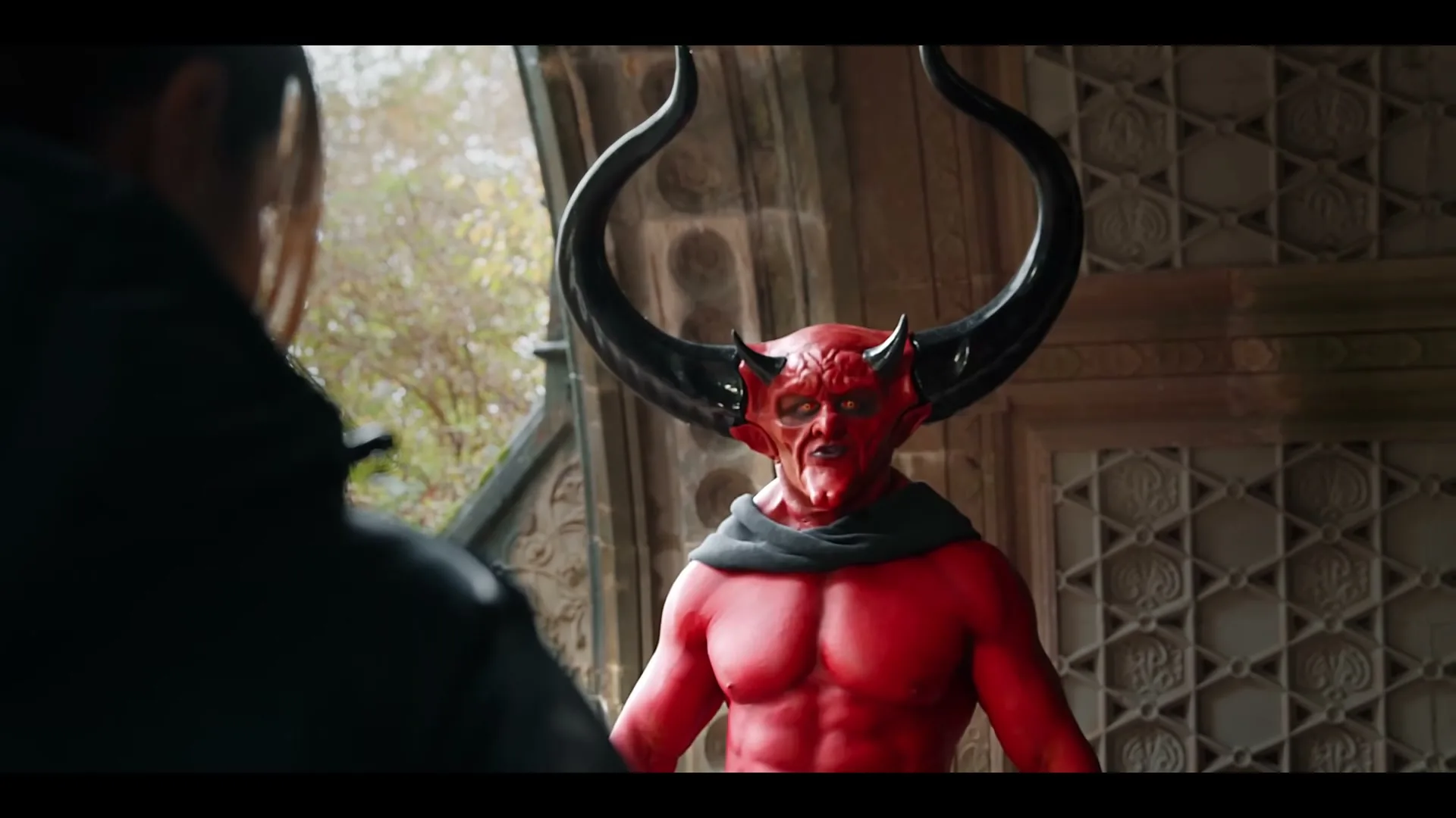 A man dressed as the Devil, with red skin and large horns, stands in a stone archway, looking at another person from behind.