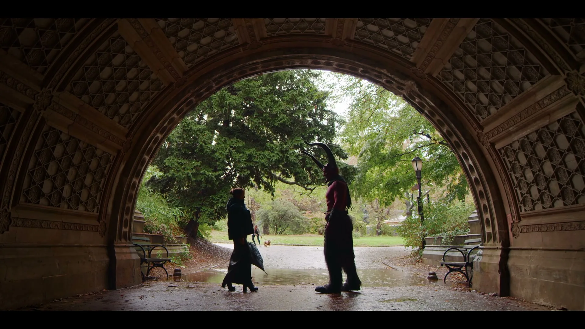 A woman in a black coat and a man dressed as the devil with horns stand facing each other under a stone archway in a park.