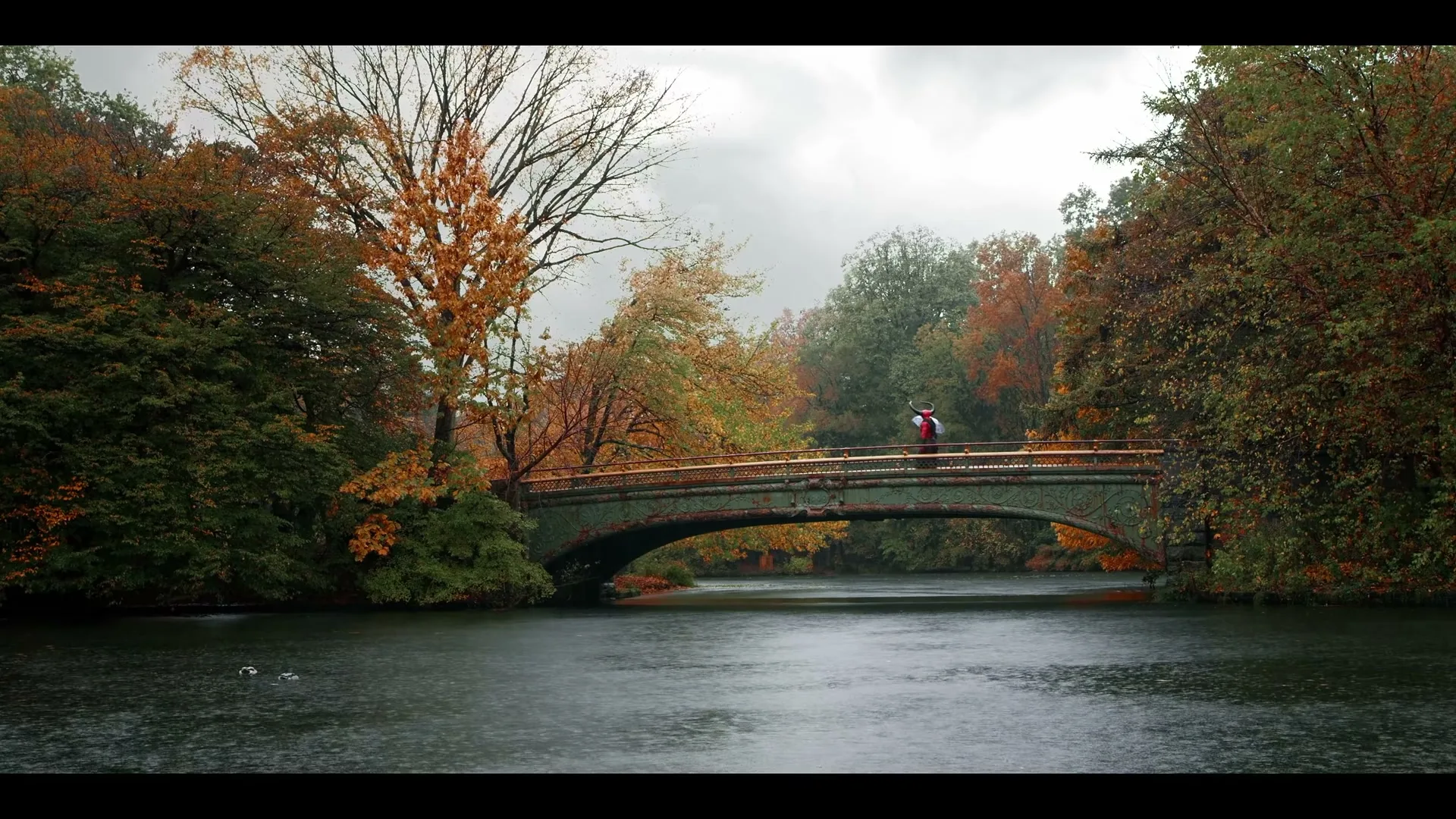 A wide shot shows Satan, a female figure with horns and a red outfit, walking across a bridge over a lake in a park during autumn.