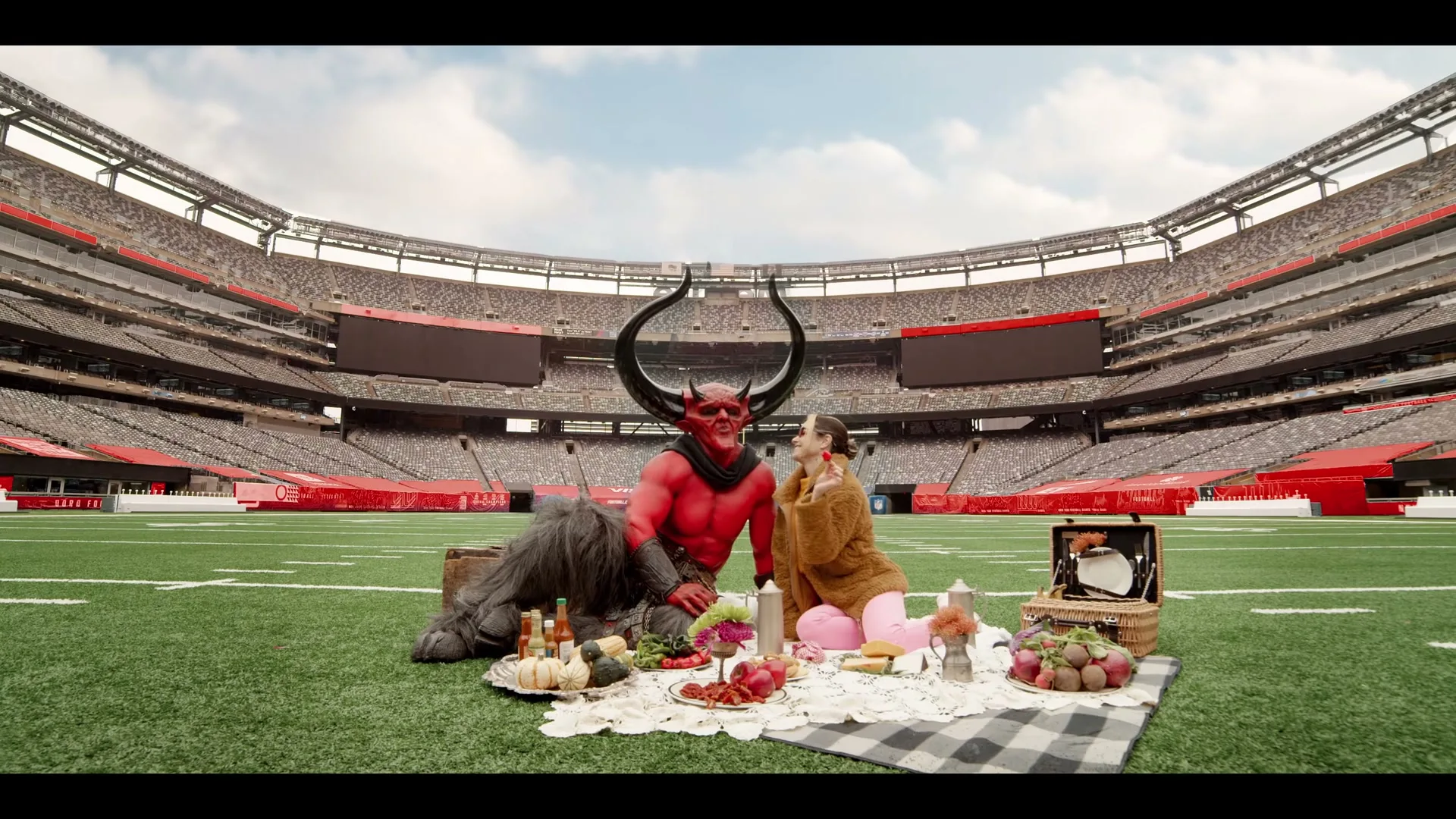 A woman in a brown coat and pink pants applies makeup to a red devil with large horns, who is sitting next to a furry creature, during a picnic on a football field.