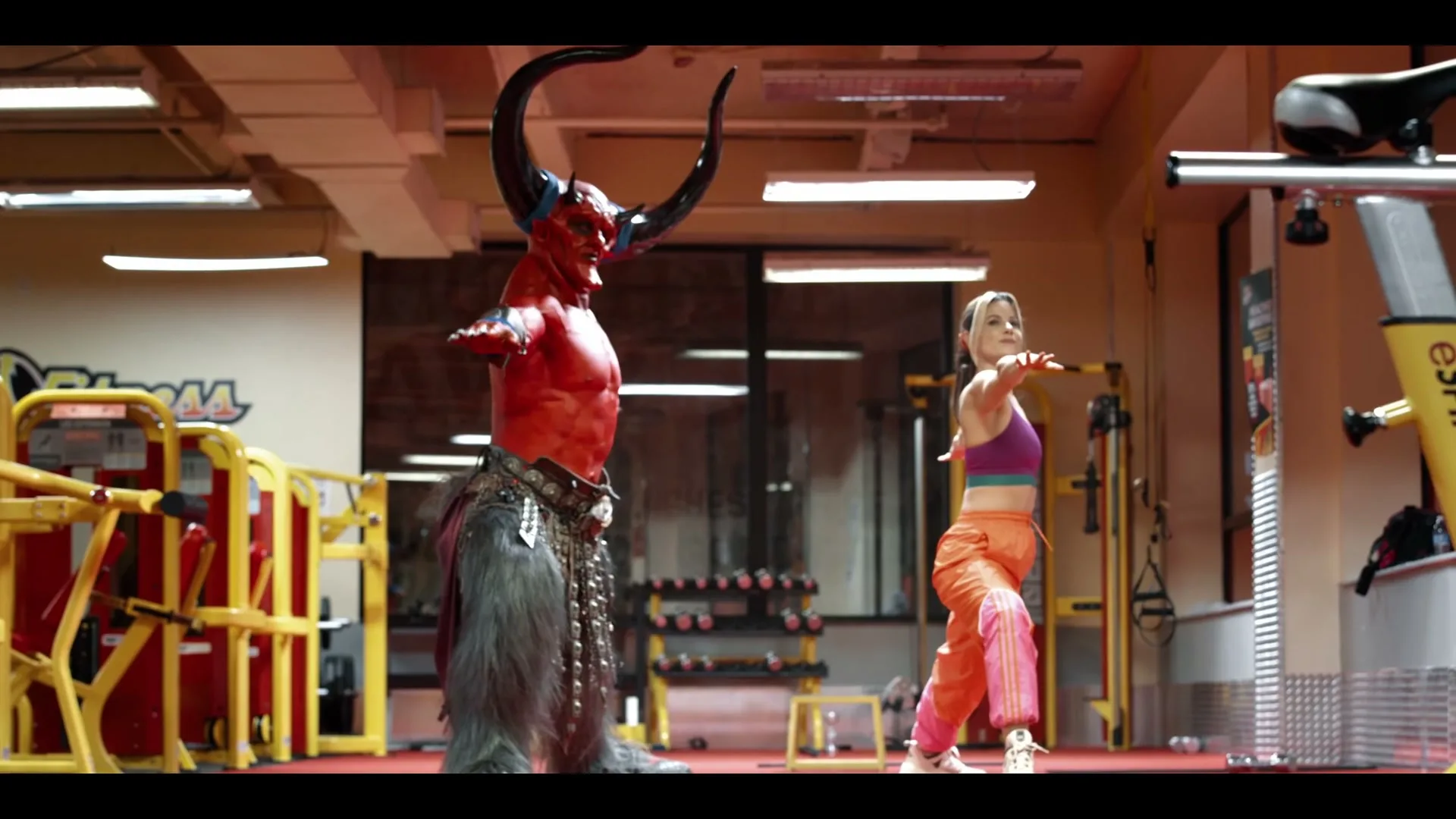A woman in workout clothes and a red devil-like creature with horns and fur do yoga poses in a brightly lit gym.