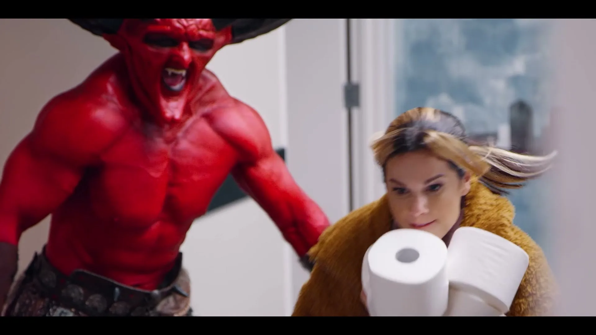 A woman in a furry coat runs away from a red devil, holding toilet paper, in a hallway with a painted door.
