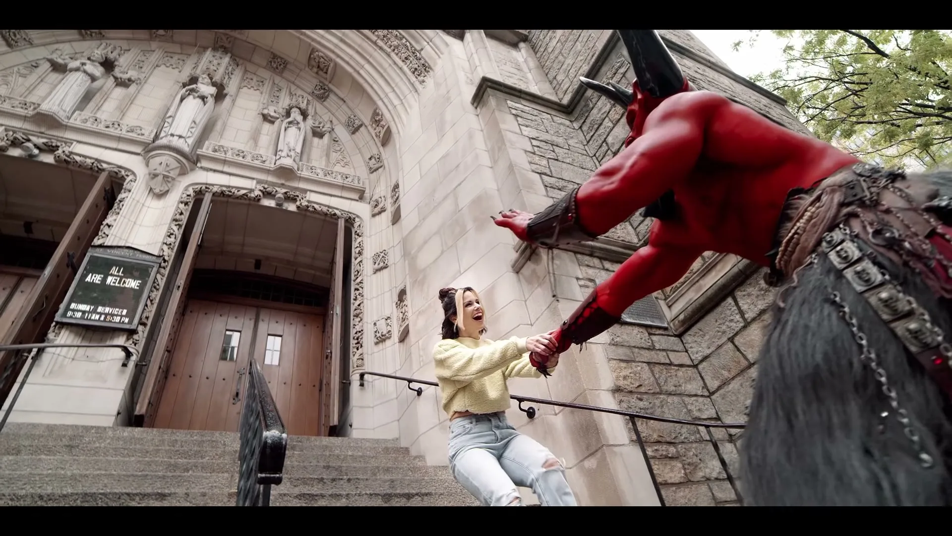A woman in a yellow sweater and ripped jeans holds hands with a red devil in front of a church, both smiling.