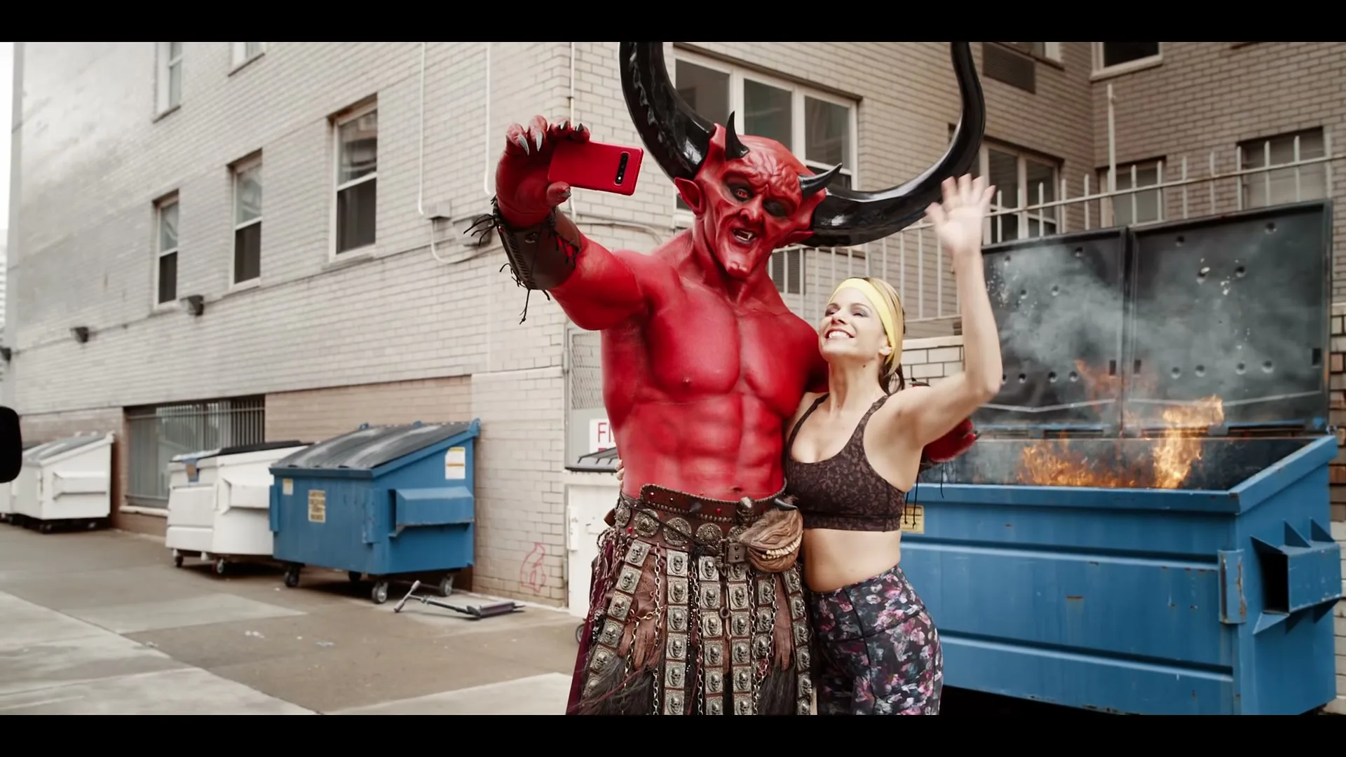 Satan, a muscular red devil with large horns, takes a selfie with a blonde woman in workout clothes in an alleyway with dumpsters, one of which is on fire.
