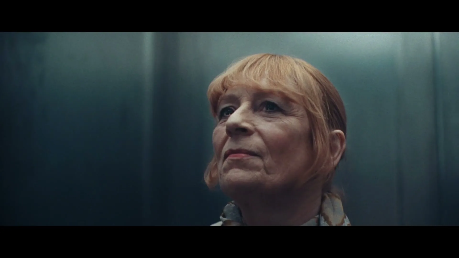 A senior woman with red hair and a patterned scarf looks up with a neutral expression inside a dimly lit elevator.