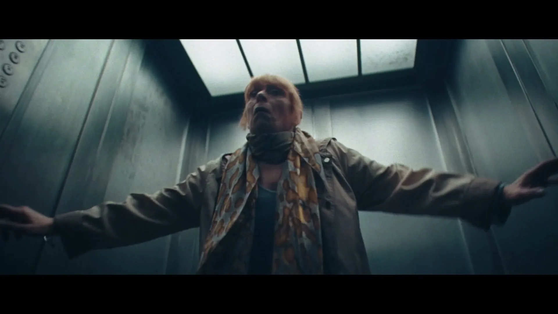 An older woman with blonde hair and a scarf stands in an elevator with her arms outstretched, looking up in fear.