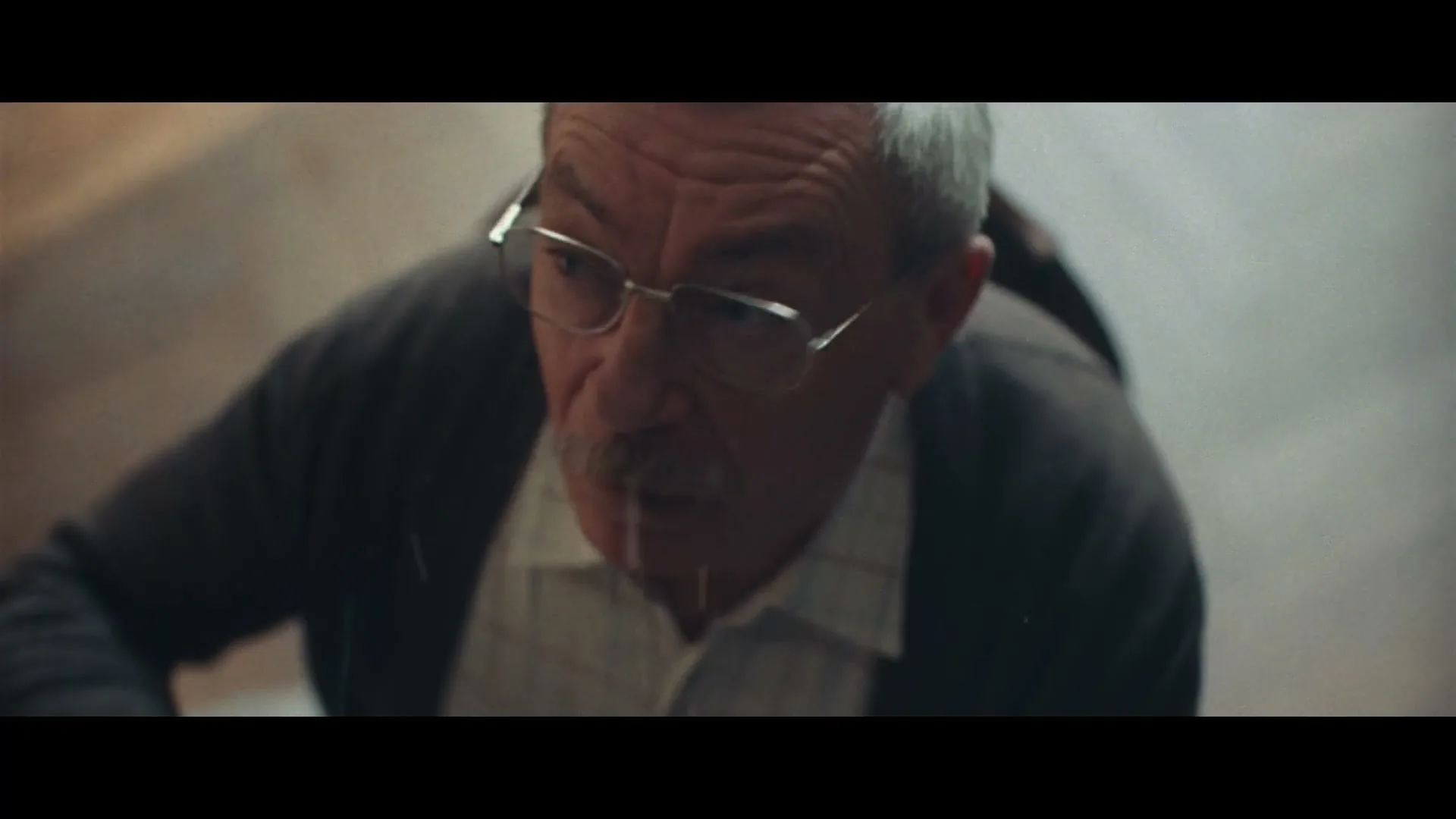 An older man with glasses and a mustache looks up with a surprised expression, as a drop of liquid falls onto his face in an interior setting.