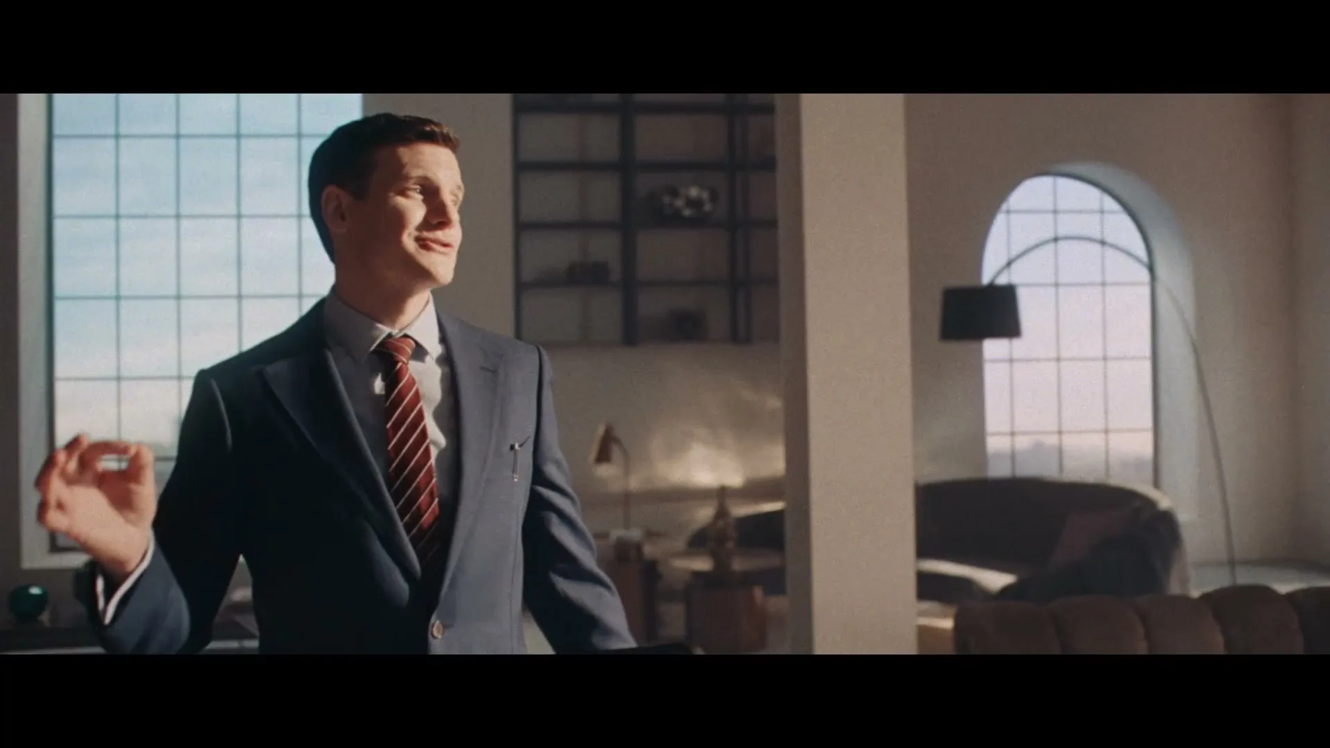 A man in a suit stands in a modern apartment with large windows, gesturing with his hand as he speaks.
