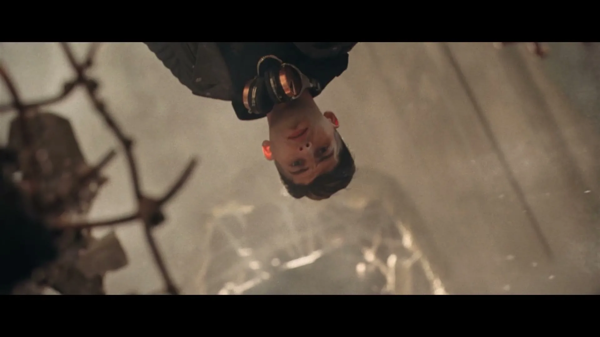 A young man with dark hair and a black jacket hangs upside down, looking at the camera with a neutral expression amidst a dusty, destroyed environment.