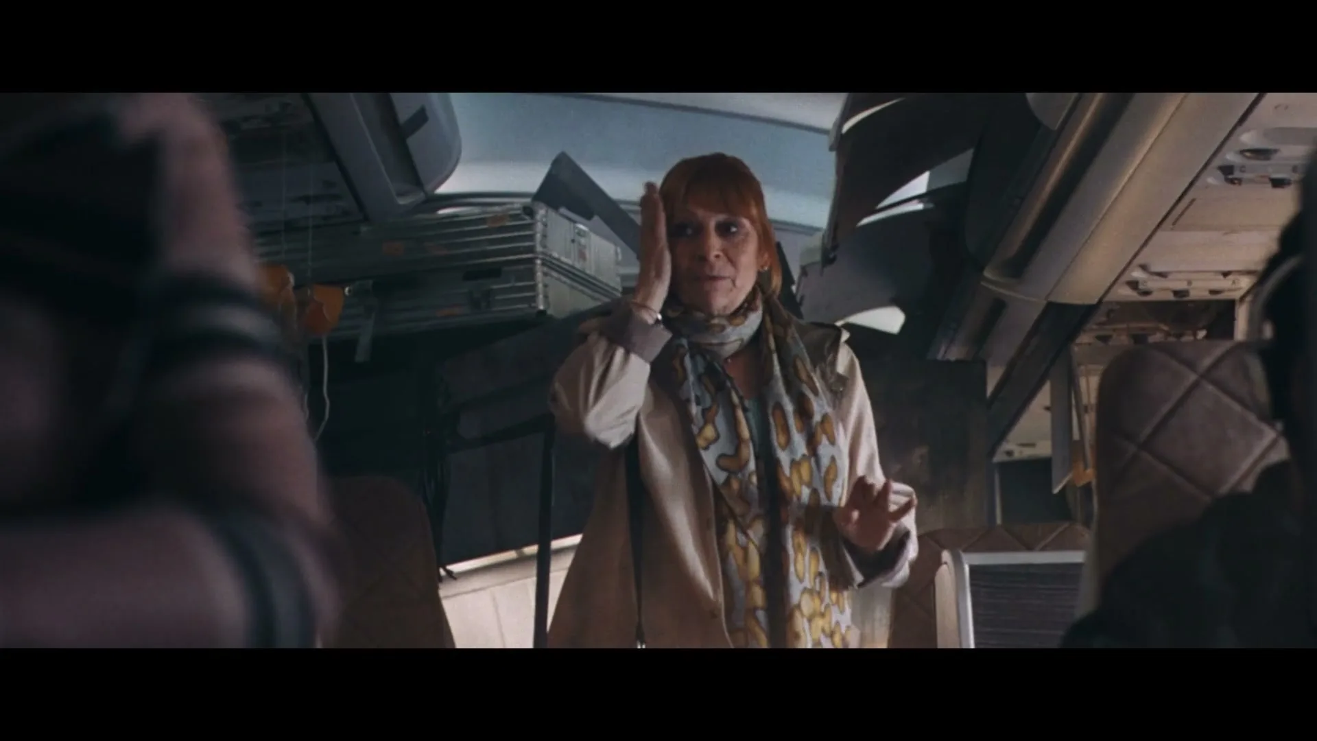 A woman with red hair and a patterned scarf stands in a damaged airplane cabin, looking distressed and holding her hand to her ear.