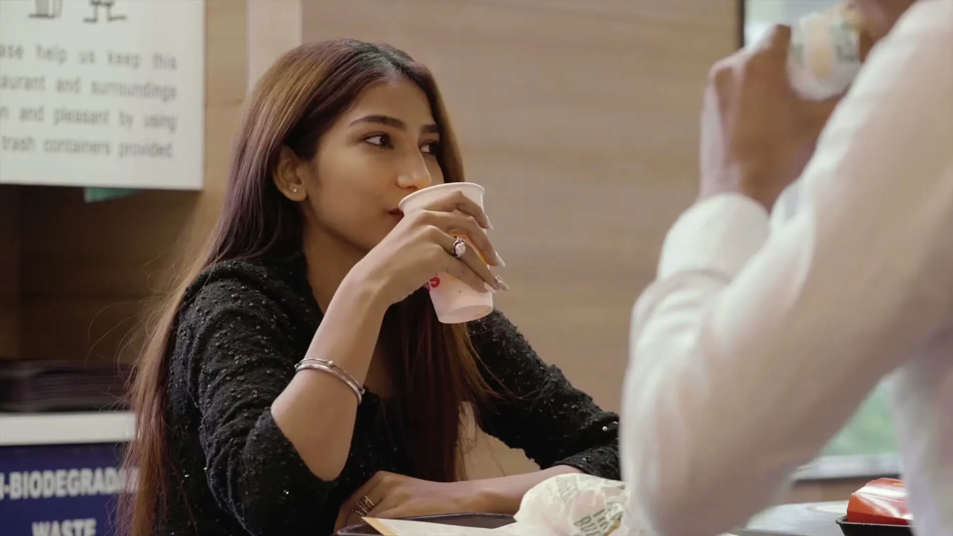 A young woman with long brown hair, wearing a black sparkly top, sips from a cup while looking to her right, sitting at a table in a McDonald's restaurant.