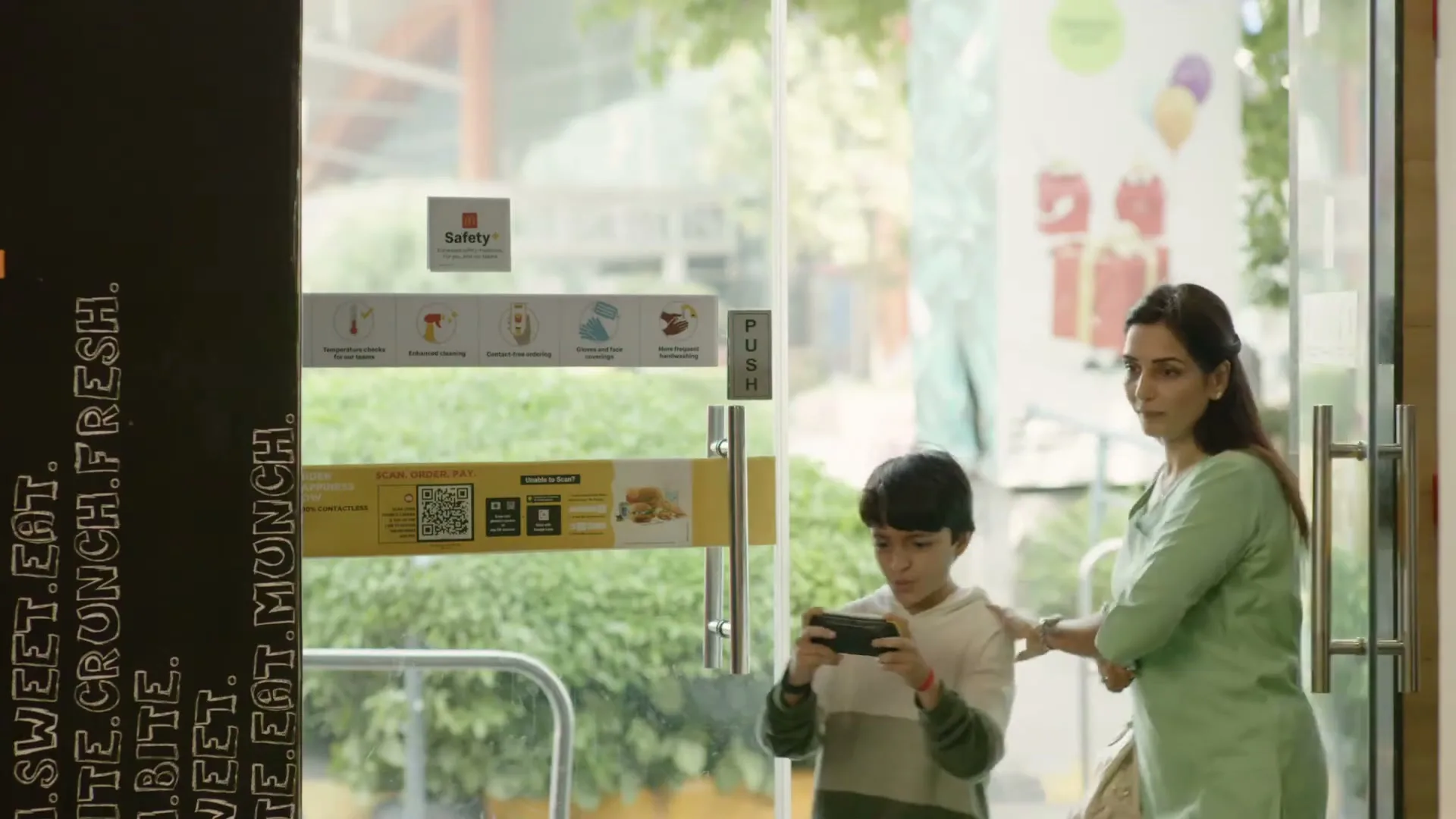 A boy in a hoodie plays a game on his phone as a woman in a green top stands next to him, entering a McDonald's restaurant.