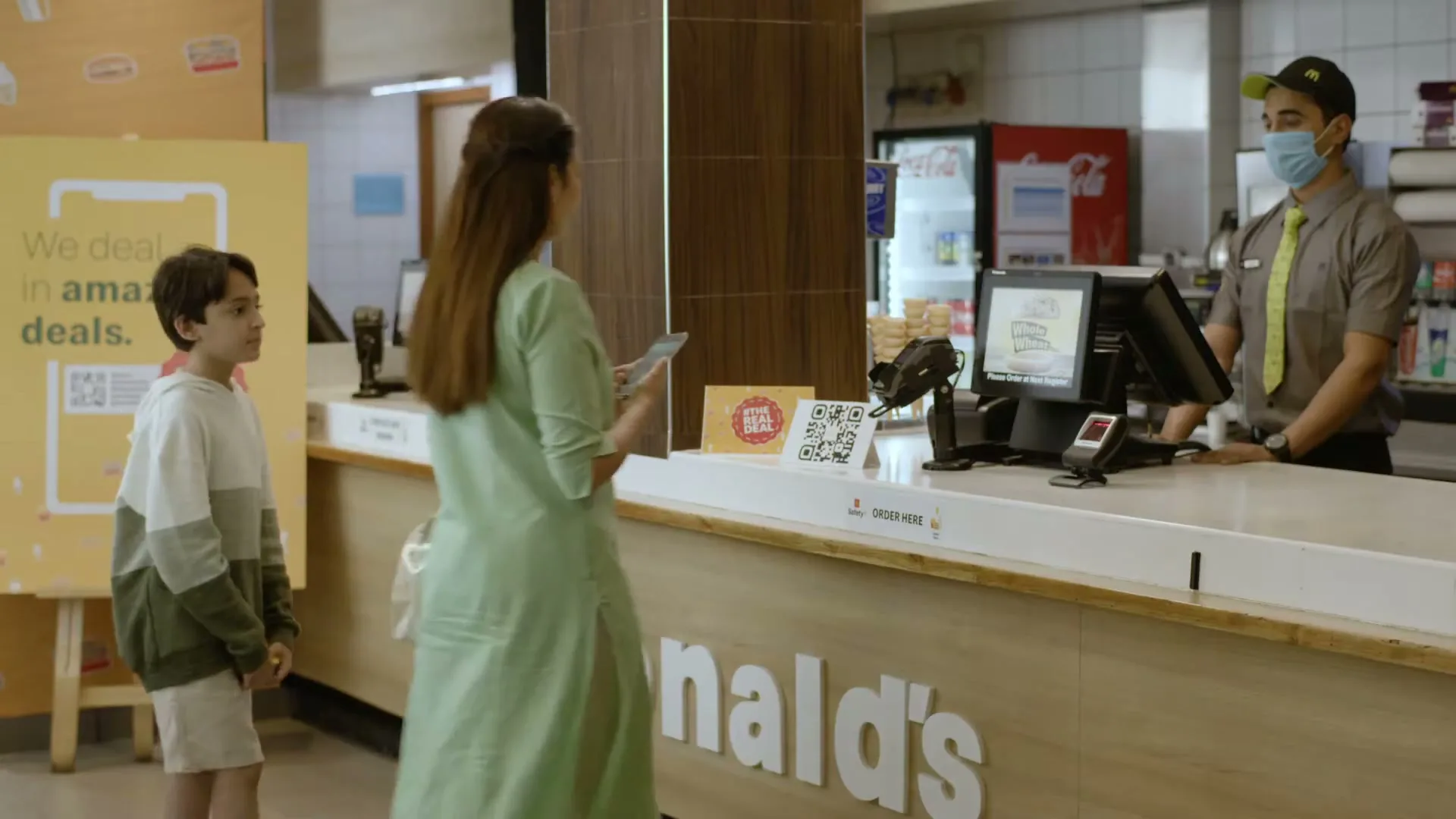 A woman and a boy stand at a McDonald's counter, looking at the menu, while a masked employee stands behind the counter.