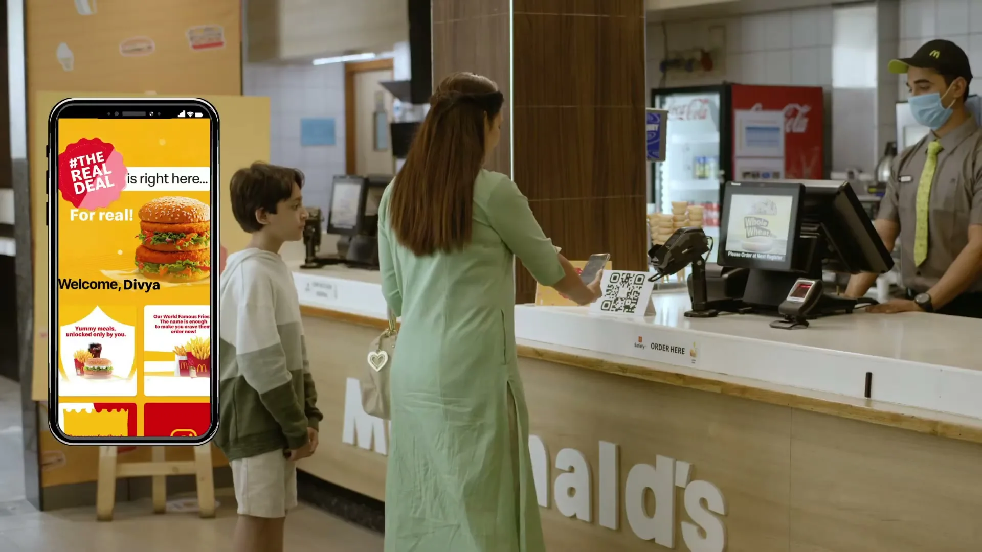 A woman and a boy stand at a McDonald's counter, while the woman scans a QR code with her phone, which displays the McDonald's app.