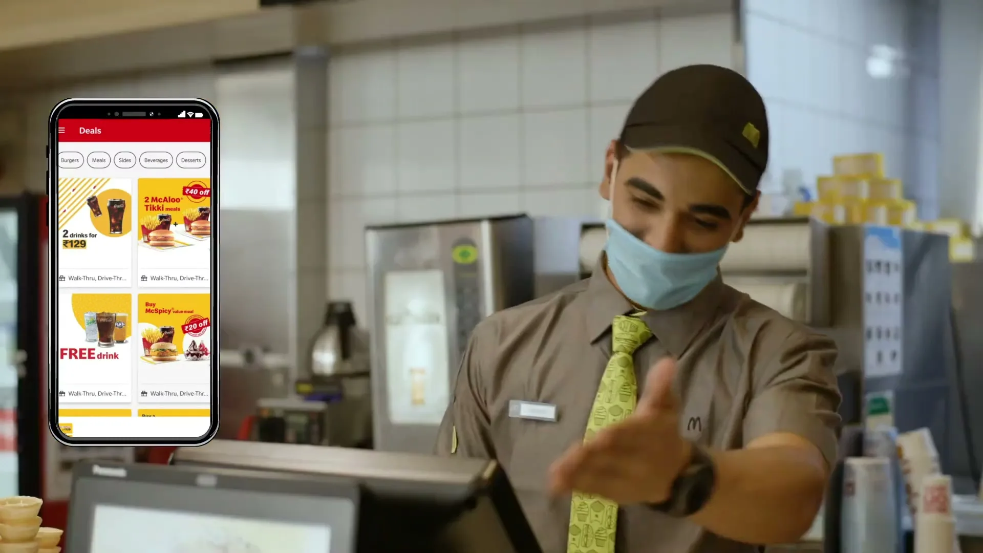 A McDonald's employee in uniform and a face mask gestures towards a phone displaying the McDonald's app with deals, inside a McDonald's restaurant.
