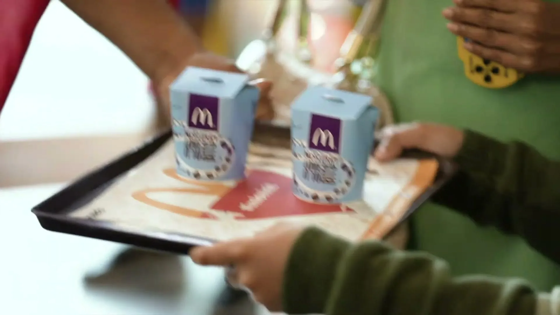 A child's hands hold a tray with two McDonald's McFlurry containers, as an adult's hands are visible in the background, in a bright, indoor setting.