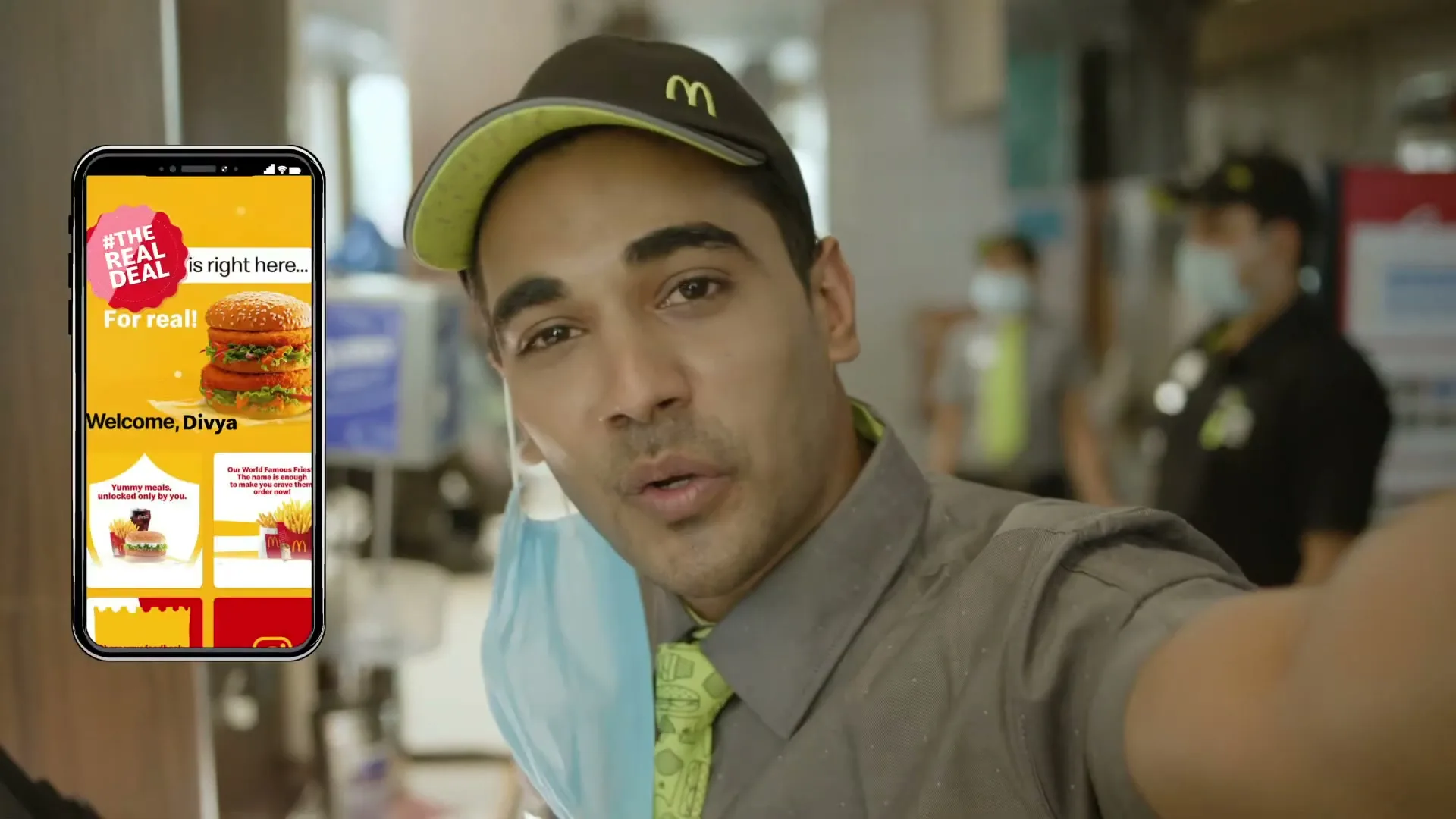 A McDonald's employee in uniform pulls down his face mask and smiles at the camera, with a phone screen showing the McDonald's app overlaid on the left.