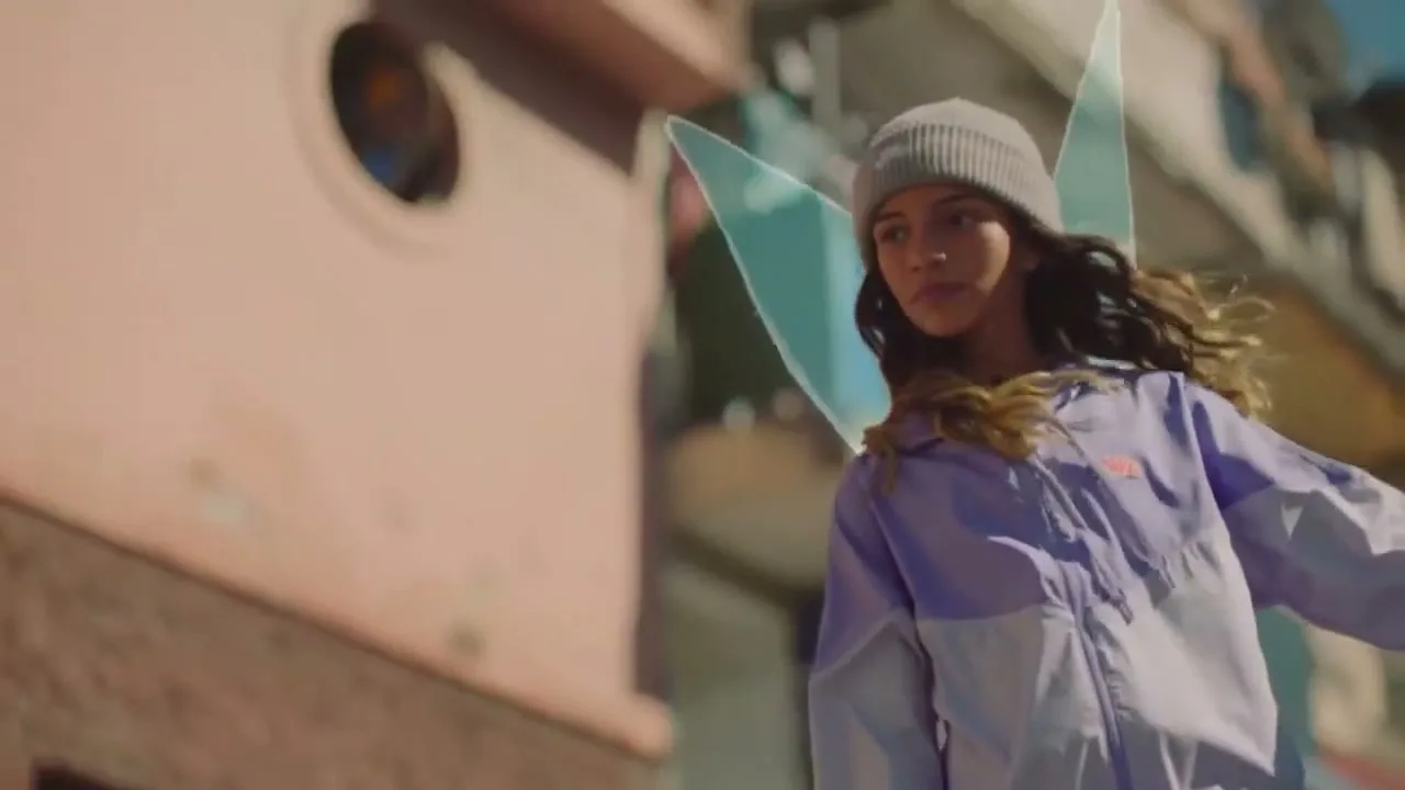 A young female with long brown hair and a beanie wears a purple and white jacket with animated fairy wings, looking off-camera in an urban setting.